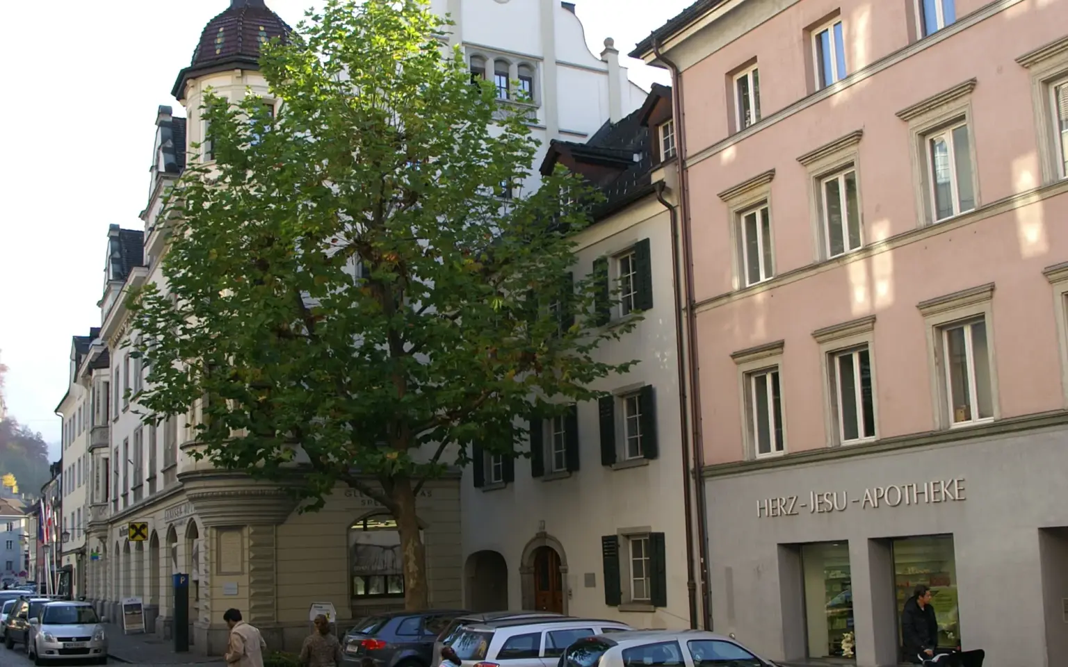 Domplatz in Feldkirch mit Platane, Turm und dem rosa Eckhaus mit dem Schriftzug „Herz-Jesu-Apotheke“ über den Schaufenstern
