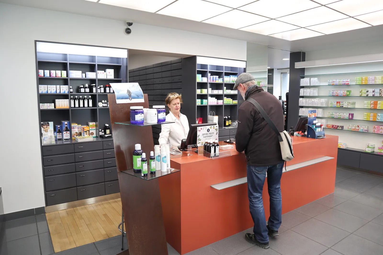 Verkaufsraum der Herz Jesu Apotheke mit kupferroter Tara, eine Apothekerin berät einen Kunden, dahinter Regale mit Arzneimitteln